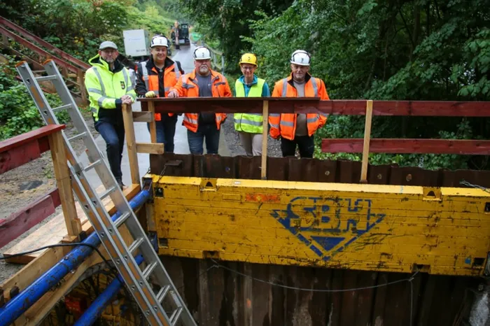Fünf Personen in Warnwesten und teilweise mit Schutzhelmen stehen an einer Baustelle für einen Stollenvortrieb hinter einem Holzgeländer.
