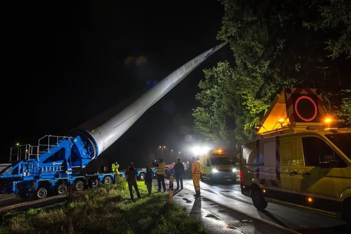 Ein großer Windradflügel wird nachts auf einem blauen Tieflader transportiert, begleitet von Personen und gelben Begleitfahrzeugen.