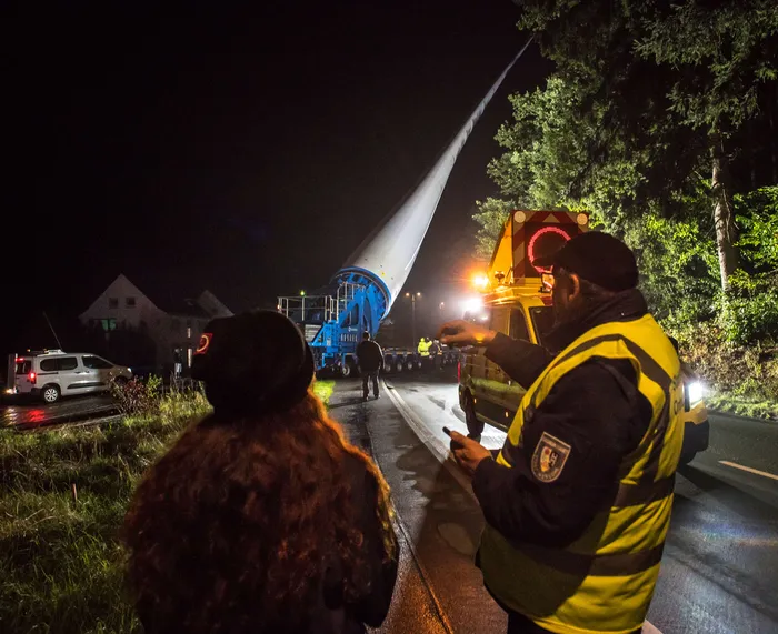 Nächtlicher Transport eines großen Windradflügels auf einem Tieflader auf einer Straße, begleitet von Personen am Straßenrand.
