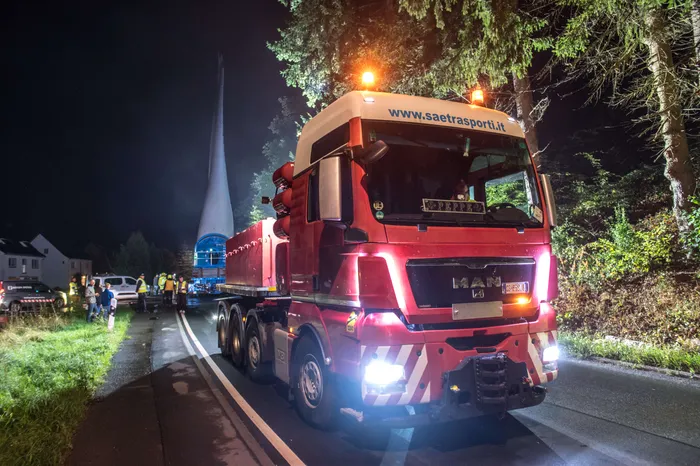 Ein roter LKW transportiert nachts einen großen, weißen Windkraftflügel auf einer Straße.