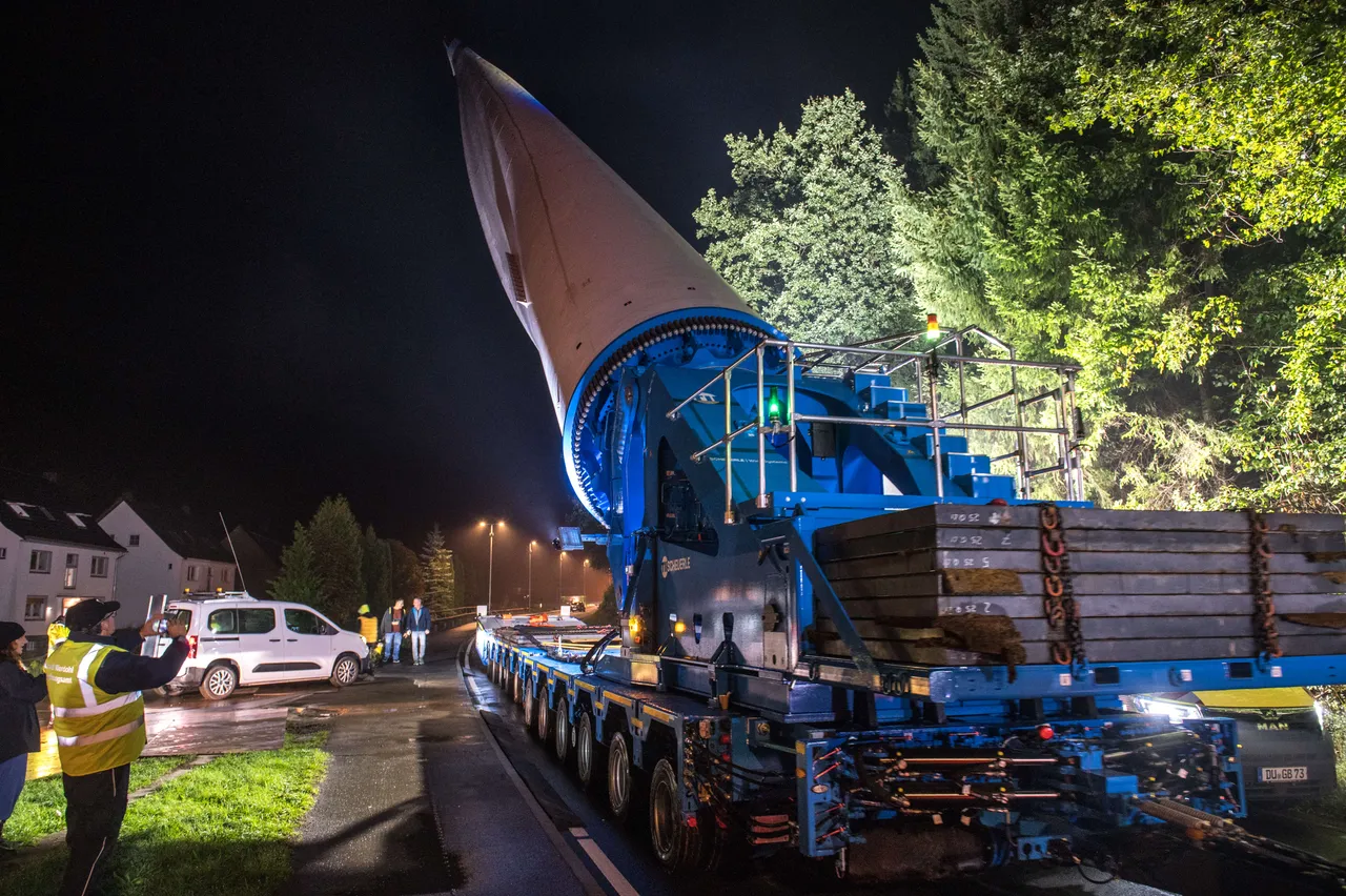Ein großer Windkraftflügel wird nachts auf einem Schwerlasttransporter transportiert, begleitet von Personal in Warnwesten.