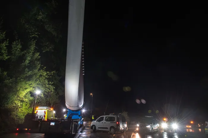 Ein großer weißer Windradflügel wird nachts auf einem Tieflader über eine Straße transportiert.