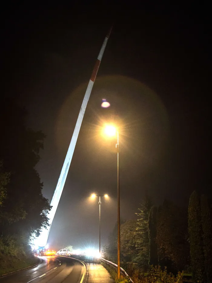 Ein langer Windradflügel wird nachts auf einer Straße transportiert und ragt steil in den dunklen Himmel.