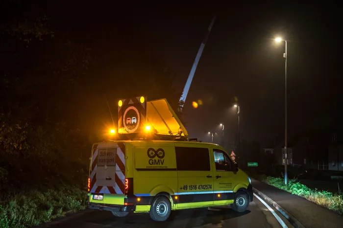 Gelber GMV-Schwerlasttransporter mit Warnleuchten und einem großen weißen Ausleger auf einer nächtlichen Straße.