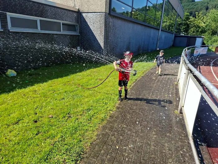 Ein Kind in roter Sportkleidung bespritzt mit einem Wasserschlauch andere Kinder auf einem Sportplatz vor einem grauen Gebäude. Im Hintergrund ist ein Handballfeld zu erkennen.