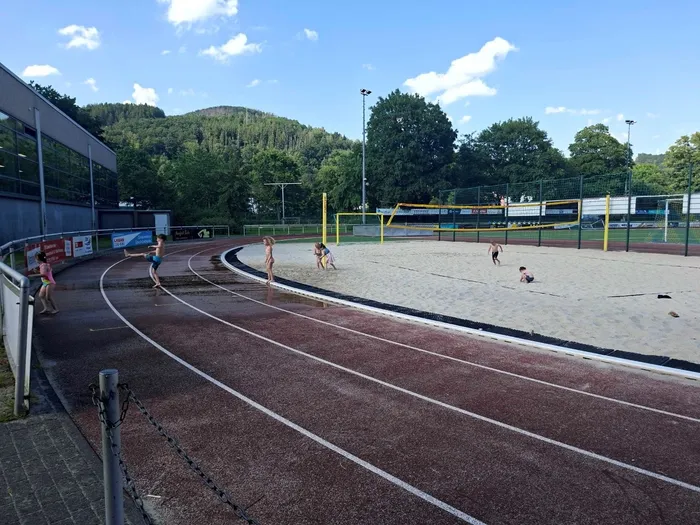 Kinder planschen auf einer Sandfläche neben einer roten Tartanbahn, während im Hintergrund ein Handballfeld zu sehen ist. Im Hintergrund sind Gebäude und Bäume erkennbar.