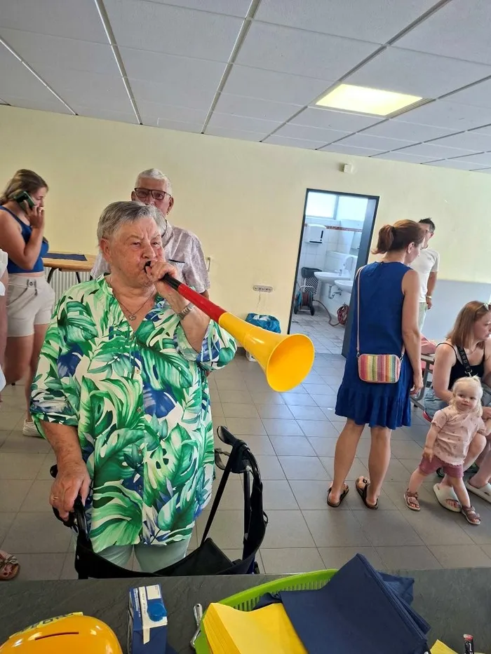 Eine ältere Frau mit kurzem, grauem Haar spielt auf einer gelb-orangen Tröte, während sie in einem Rollstuhl sitzt. Im Hintergrund sind weitere Menschen zu sehen, darunter eine Frau mit einem Baby auf dem Arm. Der Raum scheint ein Vereinsheim oder eine Sporthalle zu sein.