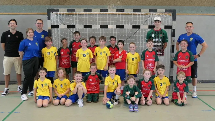 Eine Gruppe von Kindern und Jugendlichen in Handballtrikots posiert mit ihren Trainern vor einem Handballtor. Die Trikots sind in den Farben Gelb, Rot, Grün und Blau gehalten, einige tragen die Nummerierung von 2 bis 17.