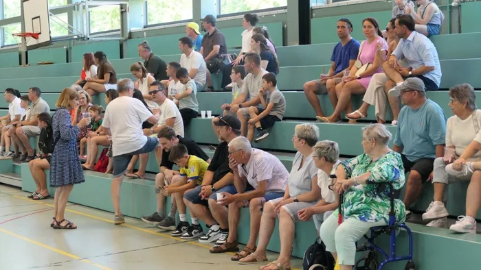 Zuschauer sitzen auf den Tribünen einer Sporthalle und beobachten ein Handballspiel. Im Vordergrund sind Kinder und Erwachsene zu sehen, einige mit Getränken oder Snacks.