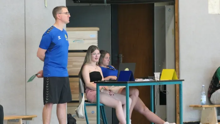 Ein Mann in einem blauen Trikot steht seitlich zu einer Frau, die an einem Tisch mit einem Laptop sitzt. Im Hintergrund sind weitere Personen und ein Tisch mit einer Wasserflasche zu sehen.