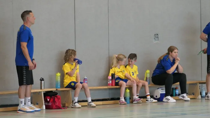 Trainer und junge Handballspielerinnen sitzen auf einer Bank in einer Sporthalle. Die Kinder in gelben Trikots trinken und lauschen ihrem Trainer.