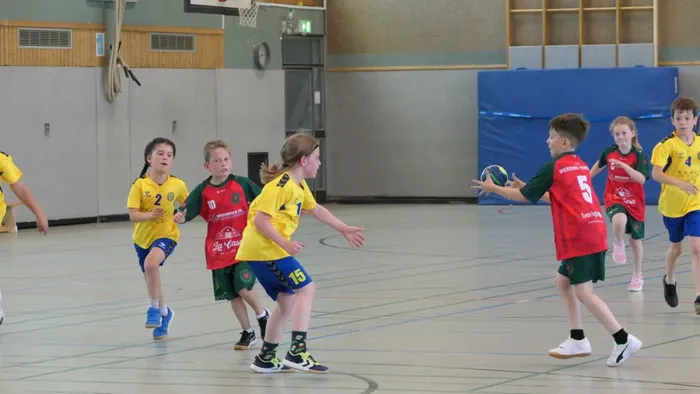 Spieler in Handballbekleidung kämpfen in einer Sporthalle um den Ball. Ein Spieler in grüner und roter Kleidung wirft den Ball, während andere Spieler in gelben Trikots versuchen, ihn abzufangen.