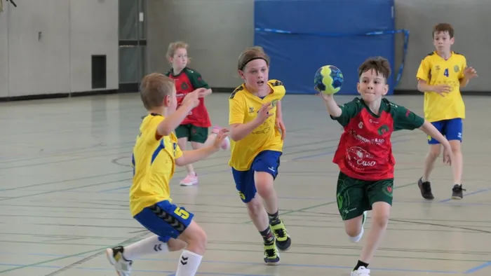 Junge Handballspieler in Aktion während eines Spiels in einer Sporthalle. Ein Spieler in Rot und Grün dribbelt den Ball, während ein Spieler in Gelb und Blau versucht, ihn zu blockieren. Im Hintergrund weitere Spieler und Zuschauer.