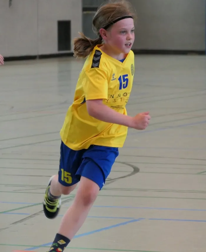 Ein Mädchen mit blonden Zöpfen und Stirnband rennt in einem gelben Handballtrikot mit der Nummer 15 auf dem Spielfeld. Im Hintergrund ist eine Sporthalle zu sehen.