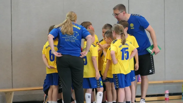 Trainerin und Trainer besprechen mit der Handball-E-Jugend des TuS Plettenberg in gelben Trikots die Taktik. Die Kinder stehen im Kreis und hören aufmerksam zu.