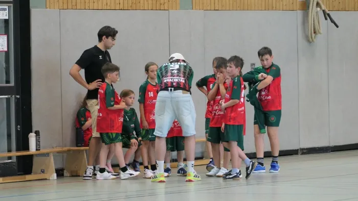 Ein Trainer steht vor einer Gruppe von Kindern in Handballkleidung und spricht sie an. Die Kinder tragen rote und grüne Trikots und hören aufmerksam zu.