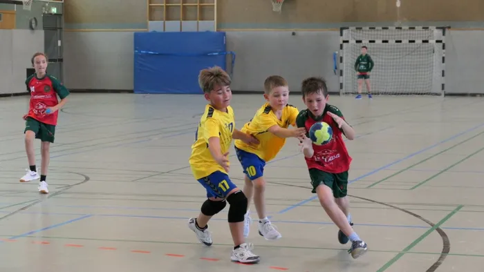 Junge Handballspieler in gelben und dunkelgrünen Trikots kämpfen um den Ball in einer Sporthalle. Im Hintergrund sind weitere Spieler und ein Handballtor zu sehen.