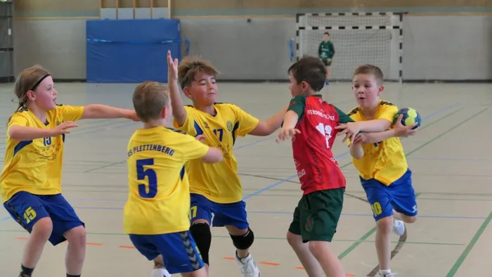 Handballspiel der E-Jugend: Zwei Spieler in gelben Trikots mit der Nummer 5 und 10 kämpfen um den Ball, während weitere Spieler in gelben, roten und blauen Trikots im Hintergrund stehen. Im Hintergrund ist ein Tor und ein Spieler in grüner Kleidung zu sehen.