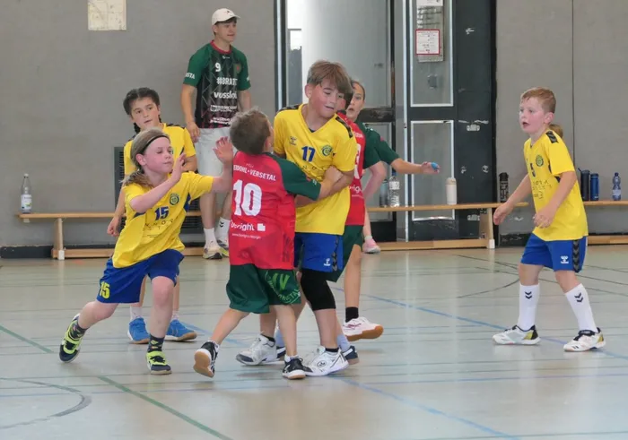Handballspiel der E-Jugend zwischen TuS Plettenberg und HSV Werdohl/Versetal in einer Sporthalle. Ein Spieler in rotem Trikot mit der Nummer 10 ist im Angriff, umringt von Verteidigern in gelben und grünen Trikots. Im Hintergrund steht ein Trainer.