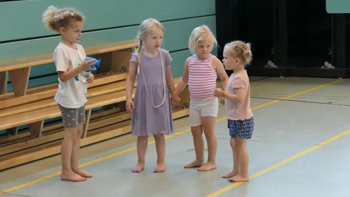 Vier junge Mädchen stehen barfuß in einer Turnhalle und halten sich an den Händen. Sie scheinen zu üben oder sich auf ein Spiel vorzubereiten. Im Hintergrund sind Turnbänke zu sehen.
