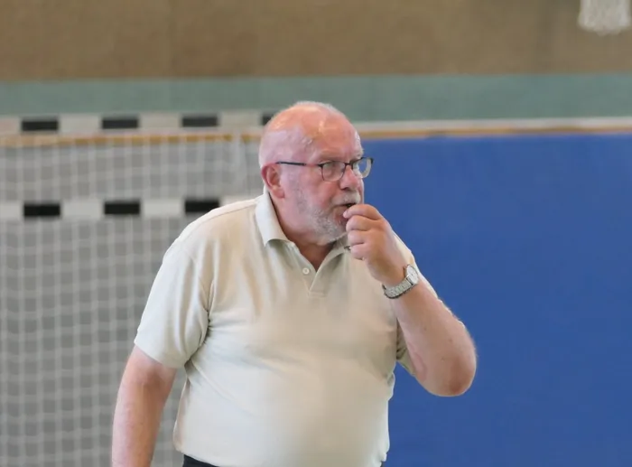 Ein älterer Mann mit Bart und Brille steht in einer Sporthalle und hält sich nachdenklich an den Mund. Im Hintergrund ist eine blaue Wand mit Markierungen zu sehen.