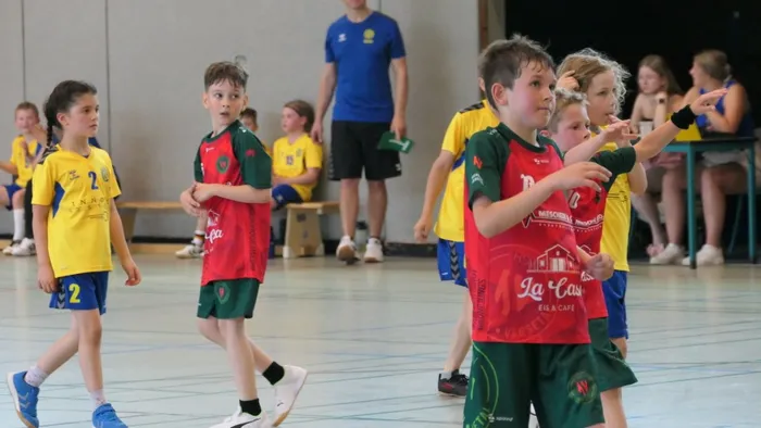 Kinder spielen Handball in einer Sporthalle. Einige tragen gelbe Trikots mit der Nummer 2, andere rote und grüne Trikots mit Werbeaufdruck. Ein Spieler zeigt mit der Hand in eine Richtung.