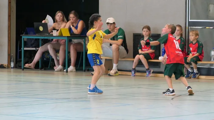 Zwei junge Handballspielerinnen stehen sich in einer Sporthalle gegenüber, während andere Kinder und Zuschauer im Hintergrund zusehen. Die Spielerin in Gelb und Blau versucht, an der Spielerin in Rot und Grün vorbeizukommen.