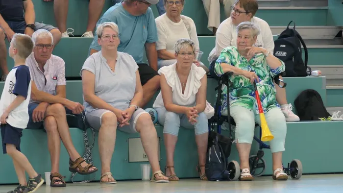 Zuschauer sitzen oder stehen an der Seitenlinie einer Sporthalle und beobachten ein Handballspiel. Eine Frau im Rollstuhl hält eine gelbe Tröte hoch.