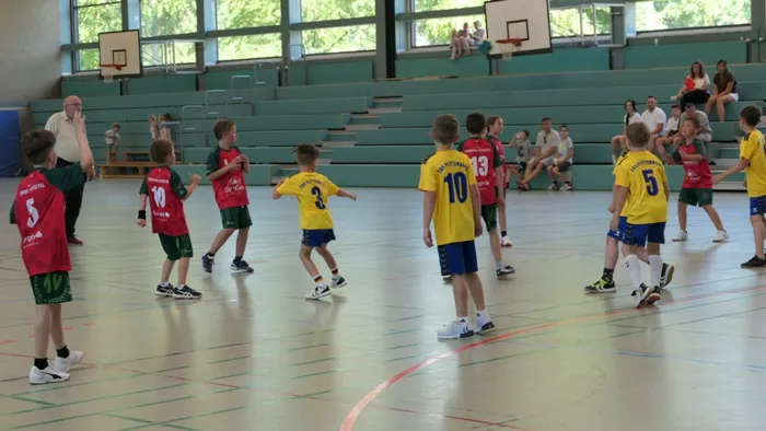 Handballspiel der E-Jugend zwischen TuS Plettenberg (rot/grün) und HSV Werdohl/Versetal (gelb/blau) in einer Sporthalle. Ein Spieler mit dem Ball im Vordergrund, weitere Spieler und Zuschauer im Hintergrund.