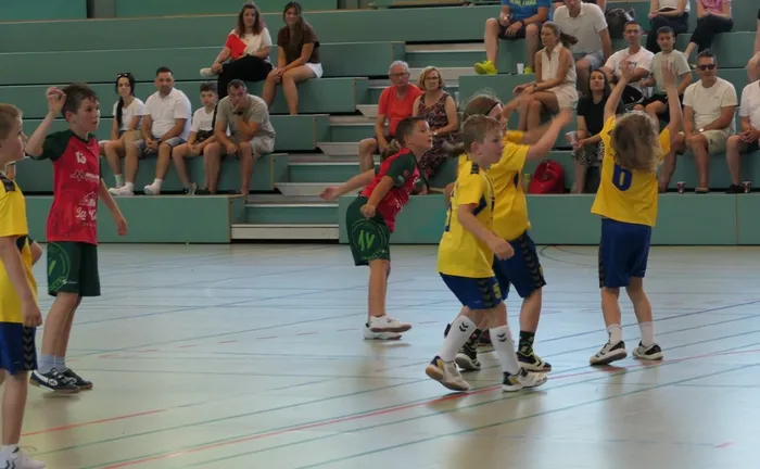Handballspiel der E-Jugend: Zwei Spieler in gelben Trikots werfen den Ball, während ein Spieler in rotem Trikot versucht, den Ball zu blocken. Im Hintergrund sitzen Zuschauer auf einer Tribüne.