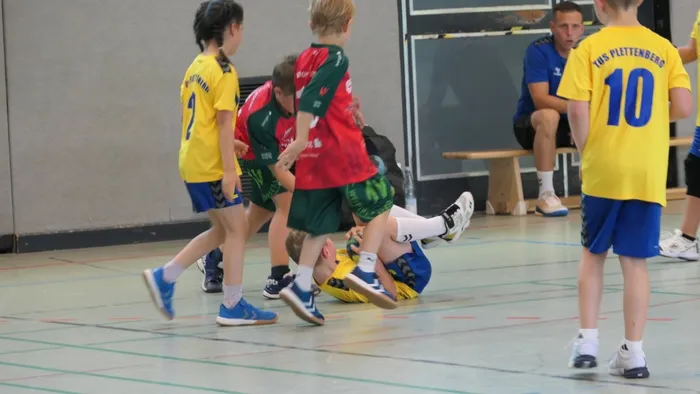 Handballspiel der E-Jugend: Ein Spieler in blauem Trikot stolpert, während andere Spieler in gelben, roten und grünen Trikots um ihn herum stehen. Im Hintergrund ein Zuschauer und eine Bank.
