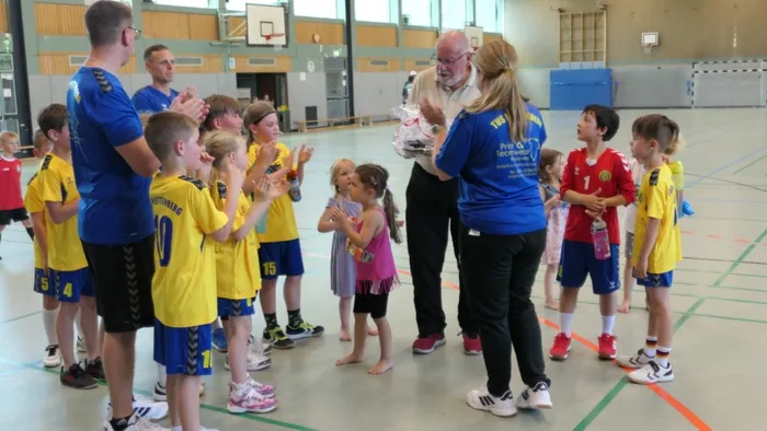 Eine Frau in blauem T-Shirt überreicht einem älteren Mann eine Blumengesteck, umringt von Kindern in Handballtrikots. Im Hintergrund stehen weitere Personen in einer Sporthalle.