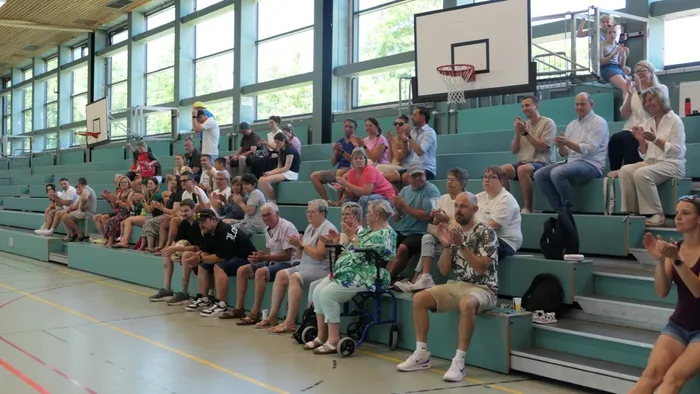 Zuschauer sitzen und stehen auf den Tribünen einer Sporthalle und applaudieren. Im Hintergrund ist ein Basketballkorb zu sehen.
