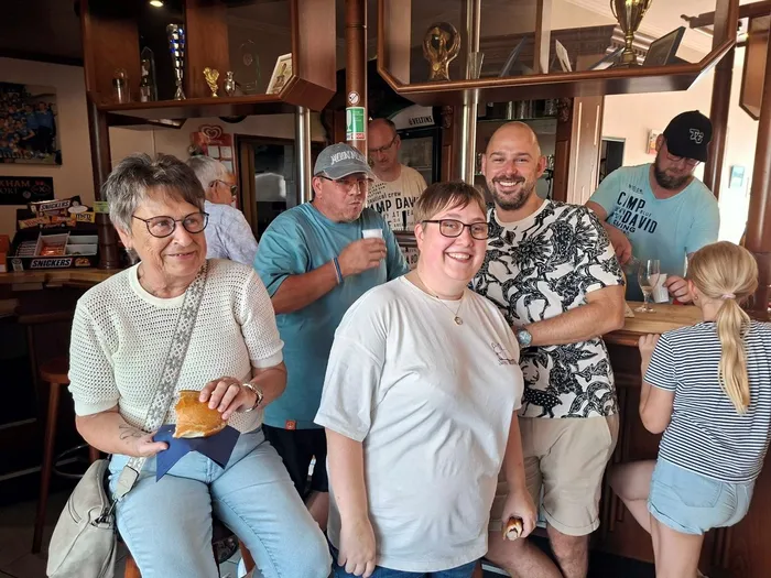 Eine Gruppe von Menschen steht und sitzt in einem Vereinsheim zusammen. Im Hintergrund sind eine Bar und Pokale zu sehen.