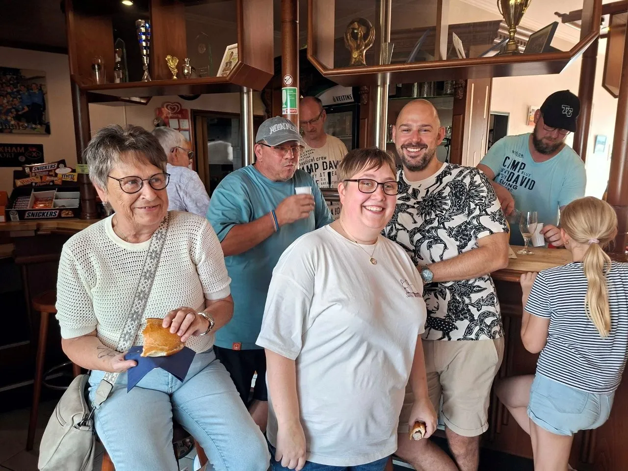 Eine Gruppe von Menschen steht und sitzt in einem Vereinsheim zusammen. Im Hintergrund sind eine Bar und Pokale zu sehen.