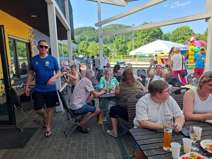 Menschen sitzen und stehen an Tischen unter einem überdachten Bereich neben einem Sportplatz, einige halten Getränke und Snacks. Im Hintergrund ist ein Fußballfeld und ein Hüpfburg zu sehen.