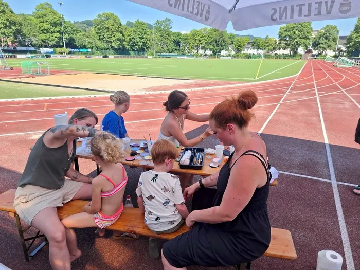 Drei Frauen sitzen an einem Tisch und bemalen die Gesichter von Kindern mit Farbe. Im Hintergrund ist eine Laufbahn und ein Sportplatz zu sehen.