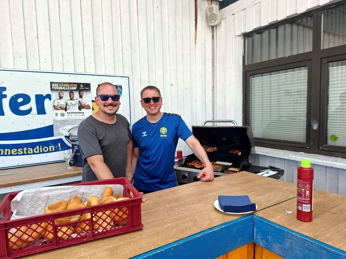 Zwei Männer stehen hinter einem Grill und bieten Würstchen an. Im Hintergrund ist ein Plakat mit Handballspielern und der Aufschrift 
