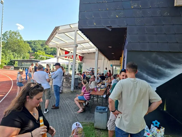 Menschen sitzen und stehen an Tischen vor einem Vereinsheim, einige halten Getränke in der Hand. Im Hintergrund ist ein Sportplatz mit einer roten Laufbahn zu sehen.