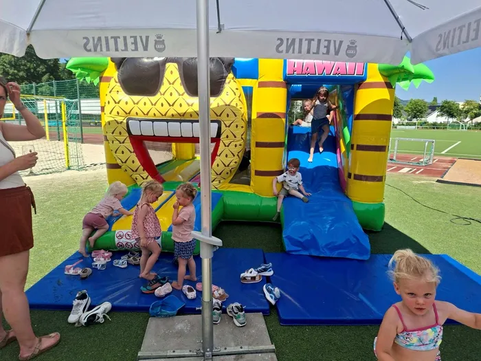 Kinder spielen auf einer bunten Hüpfburg mit Hawaii-Motiv. Im Hintergrund ist ein Sportplatz mit Laufbahn zu sehen.