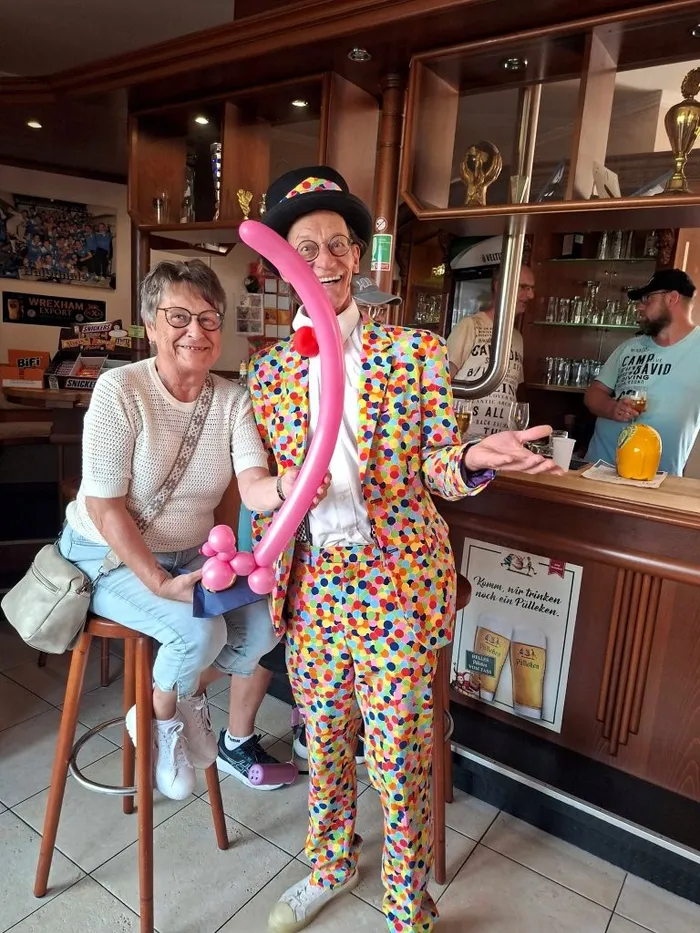 Zwei ältere Frauen, eine sitzend und eine stehend, posieren in einem Vereinsheim. Die stehende Frau trägt einen bunten Clownanzug und hält einen aufgeblasenen Luftballon. Im Hintergrund ist eine Bar mit Getränken und Dekorationen zu sehen.