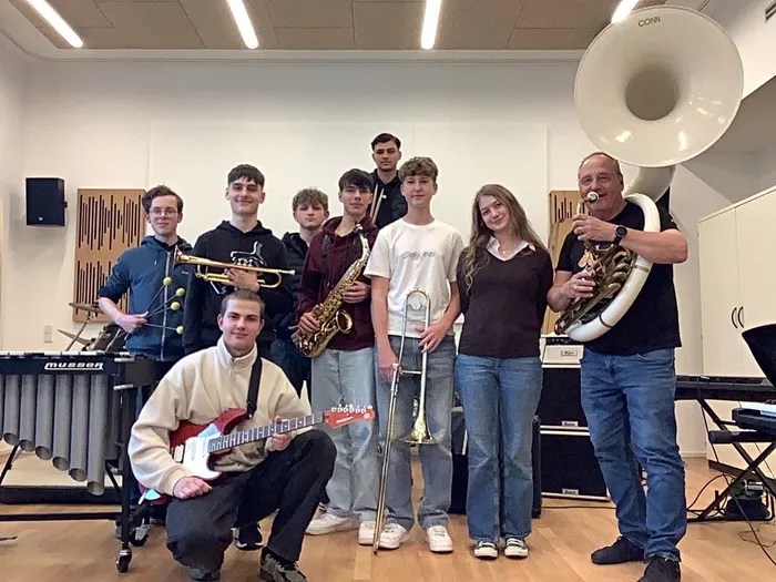 Eine Gruppe von Jugendlichen und ein Mann posieren in einem Musikraum mit Blasinstrumenten und einer Gitarre. Die Bläserbesetzung der Tuesday Jazz Band unter der Leitung von Thomas Wurth spielt Stücke aus verschiedenen Jazzrichtungen.