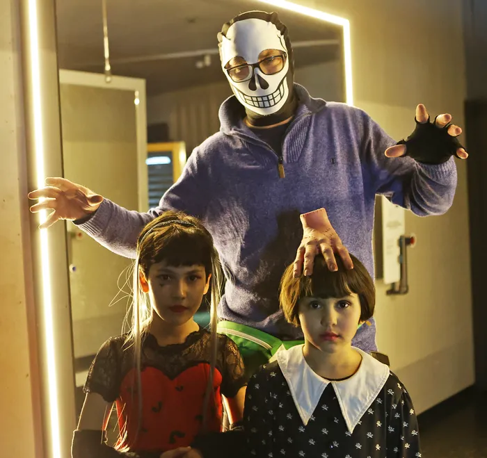 Ein Erwachsener mit Totenkopfmaske steht hinter zwei Kindern, die Kostüme tragen. Im Hintergrund ein Spiegel mit Beleuchtung.