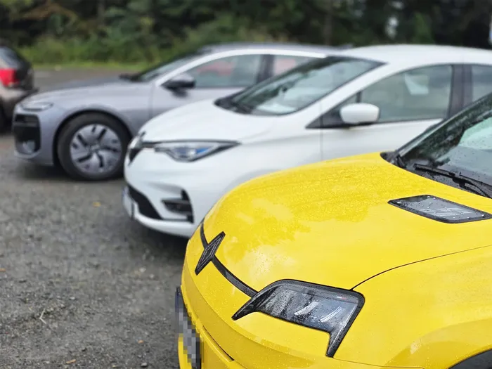 Nahaufnahme eines gelben Autos mit Wassertropfen auf der Motorhaube, dahinter stehen ein weißes und ein graues Auto auf einem Schotterplatz.