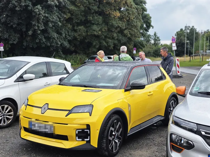 Ein gelber Renault steht im Vordergrund auf einem Parkplatz, im Hintergrund steht eine Gruppe von Menschen.