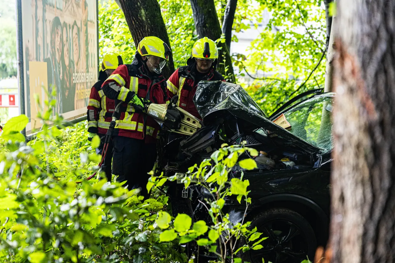 Feuerwehrleute in gelben Helmen und roten Anzügen arbeiten an einem stark beschädigten schwarzen Auto, das in Büsche und Bäume eingebettet ist. Sie sichern die Unfallstelle und trennen die Batterie ab.