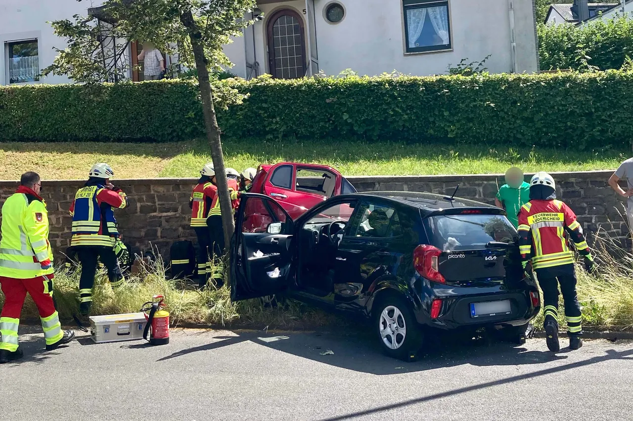 Rettungskräfte stehen um zwei beschädigte Autos, ein rotes und ein graues, nach einem Verkehrsunfall. Mehrere Einsatzkräfte in gelben Schutzanzügen und Helmen sind zu sehen.