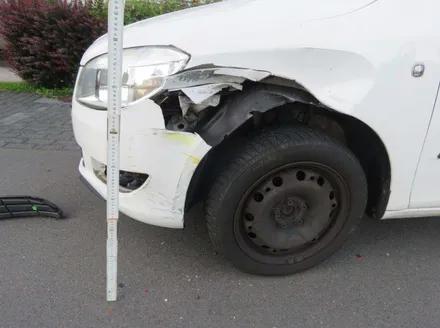 Beschädigtes weißes Auto mit stark verbeultem Kotflügel und Scheinwerfer nach einem Unfall. Ein Maßstab neben dem Schaden zeigt das Ausmaß der Beschädigung.