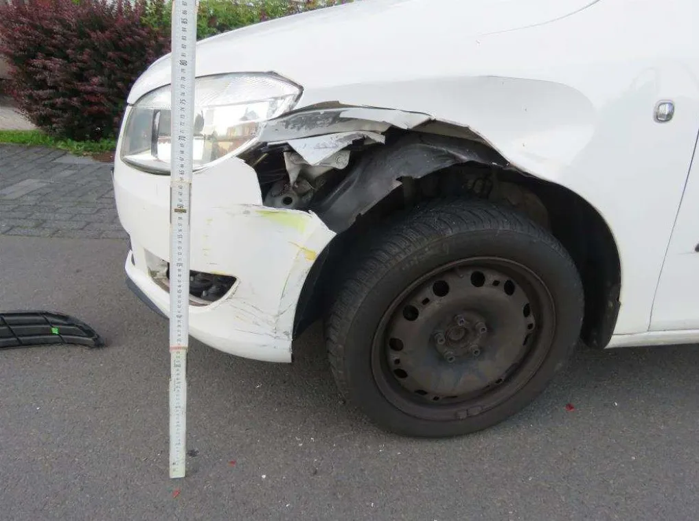 Beschädigtes weißes Auto mit stark verbeultem Kotflügel und Scheinwerfer nach einem Unfall. Ein Maßstab neben dem Schaden zeigt das Ausmaß der Beschädigung.