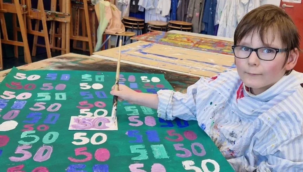 Ein Schüler mit Brille malt mit einem Pinsel auf eine große Fläche mit der Zahl 50, umgeben von farbigen Zahlen auf dem Boden.